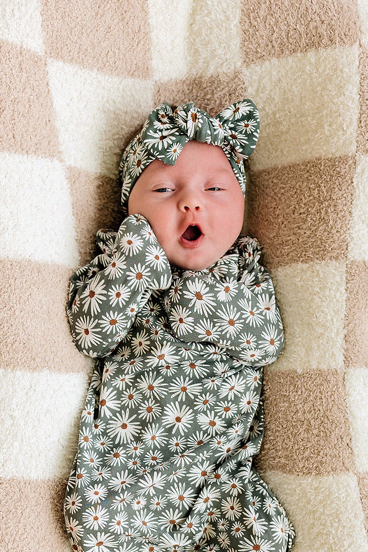 A baby wearing a dark green daisy-patterned head wrap and outfit, lying on a soft checkered blanket.