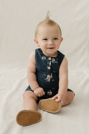 A smiling baby boy sits on a light-colored fabric background, wearing a navy cotton romper with a playful sailboat print.