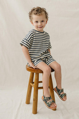 A young boy with curly hair sits on a wooden stool, wearing a navy striped shorts set and camo sandals.