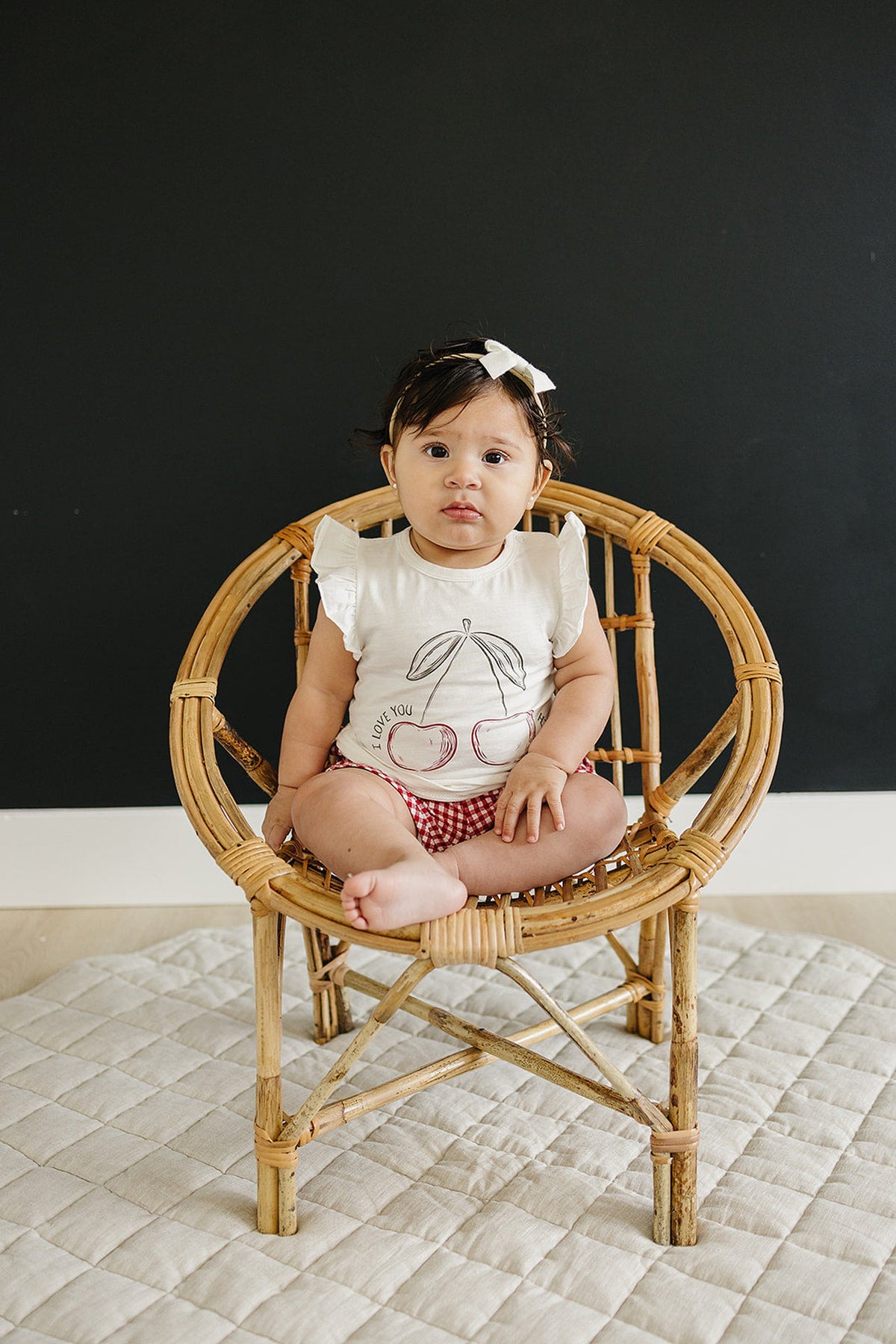 A baby wearing a white ruffle-sleeve cherry print t-shirt sits on a wicker chair against a dark background.