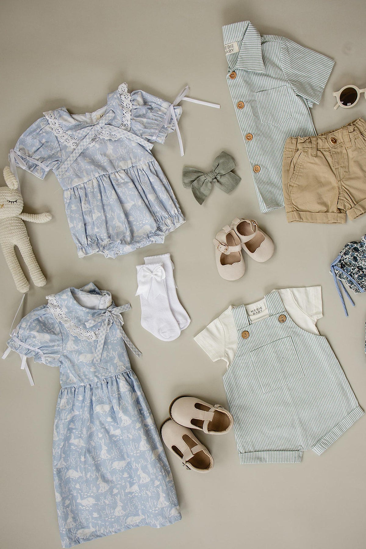 Light blue bunny-themed dress with Peter Pan collar, surrounded by baby clothes, shoes, and accessories on a beige background.
