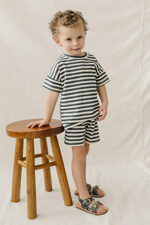 A young child poses in a navy and cream striped shorts set, standing beside a wooden stool, wearing stylish sandals.