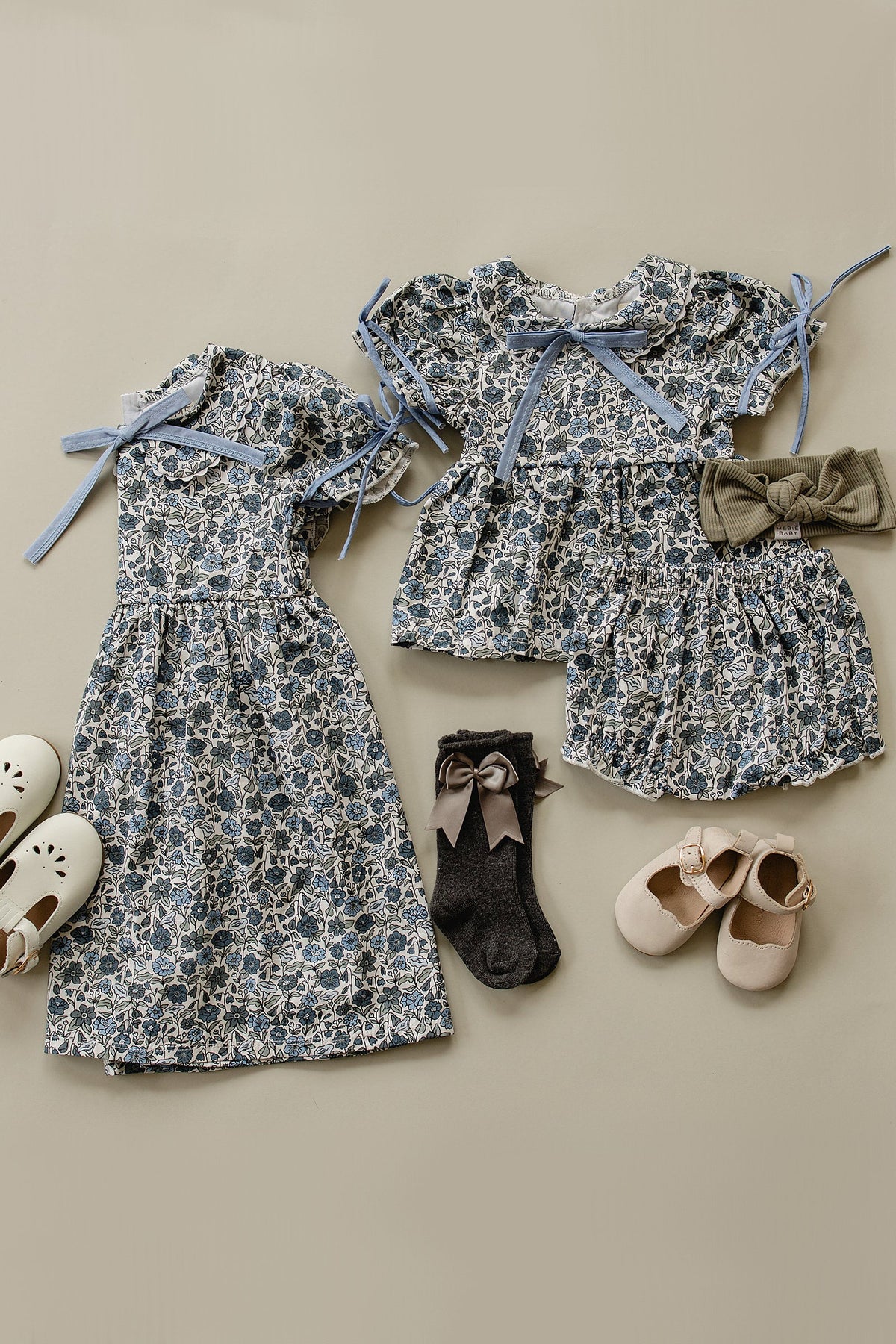 Floral-patterned baby dresses with blue bows, paired with gray bow socks, white and beige shoes, and a headband.