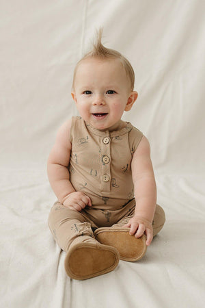 A smiling baby sits on a light-colored surface, wearing a beige tank romper with western-themed graphics and buttons.