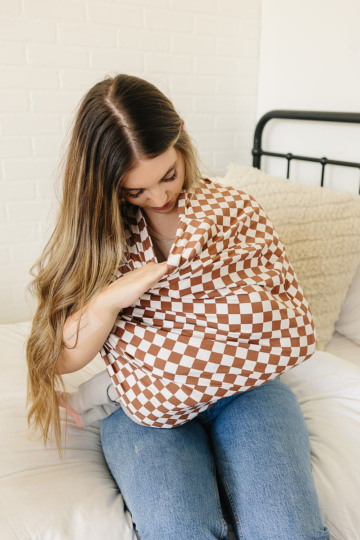 A woman sits on a bed, using a rust checkered bamboo nursing cover, with a baby underneath.