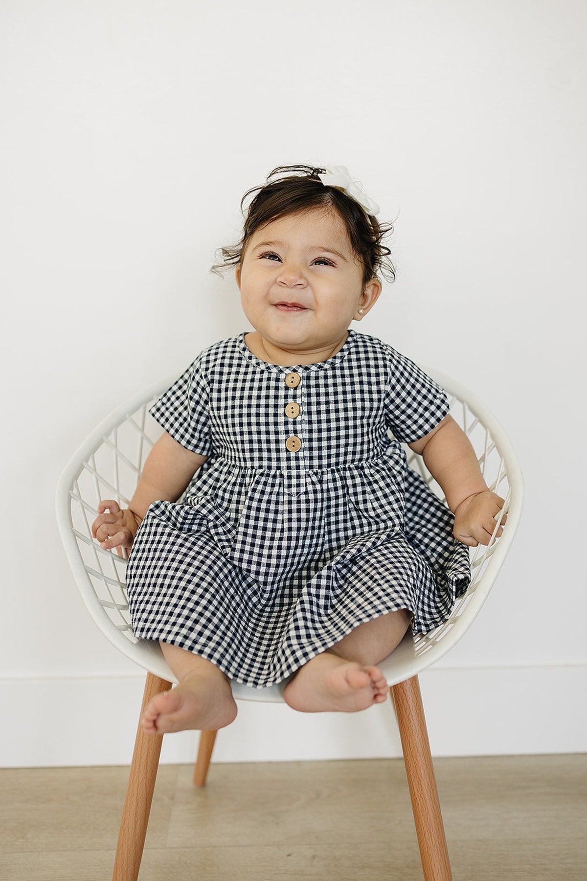 A smiling baby sits in a white chair, wearing a navy gingham cotton dress with wooden buttons.
