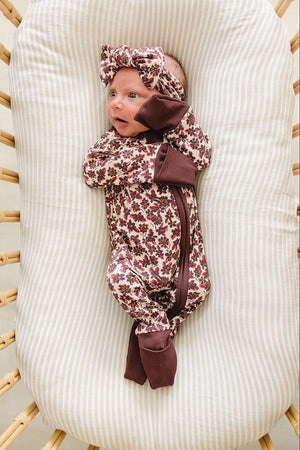 A baby lies on a soft, striped surface, wearing Antique Floral Ribbed Bamboo Zipper pajamas in burgundy and cream, complemented by a matching headband.