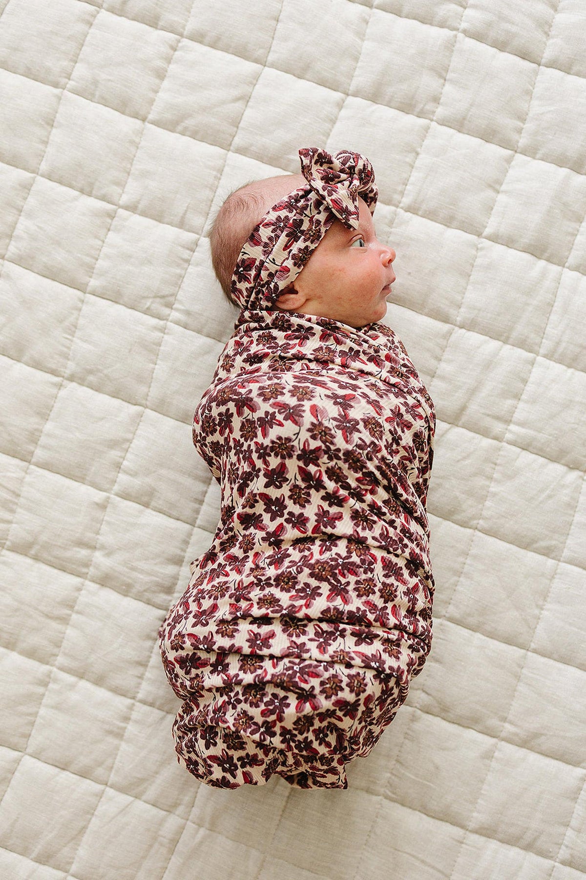 A newborn baby is swaddled in a floral ribbed bamboo stretch blanket, wearing a matching headband. The soft, breathable fabric features a delicate floral pattern against a neutral quilted background.