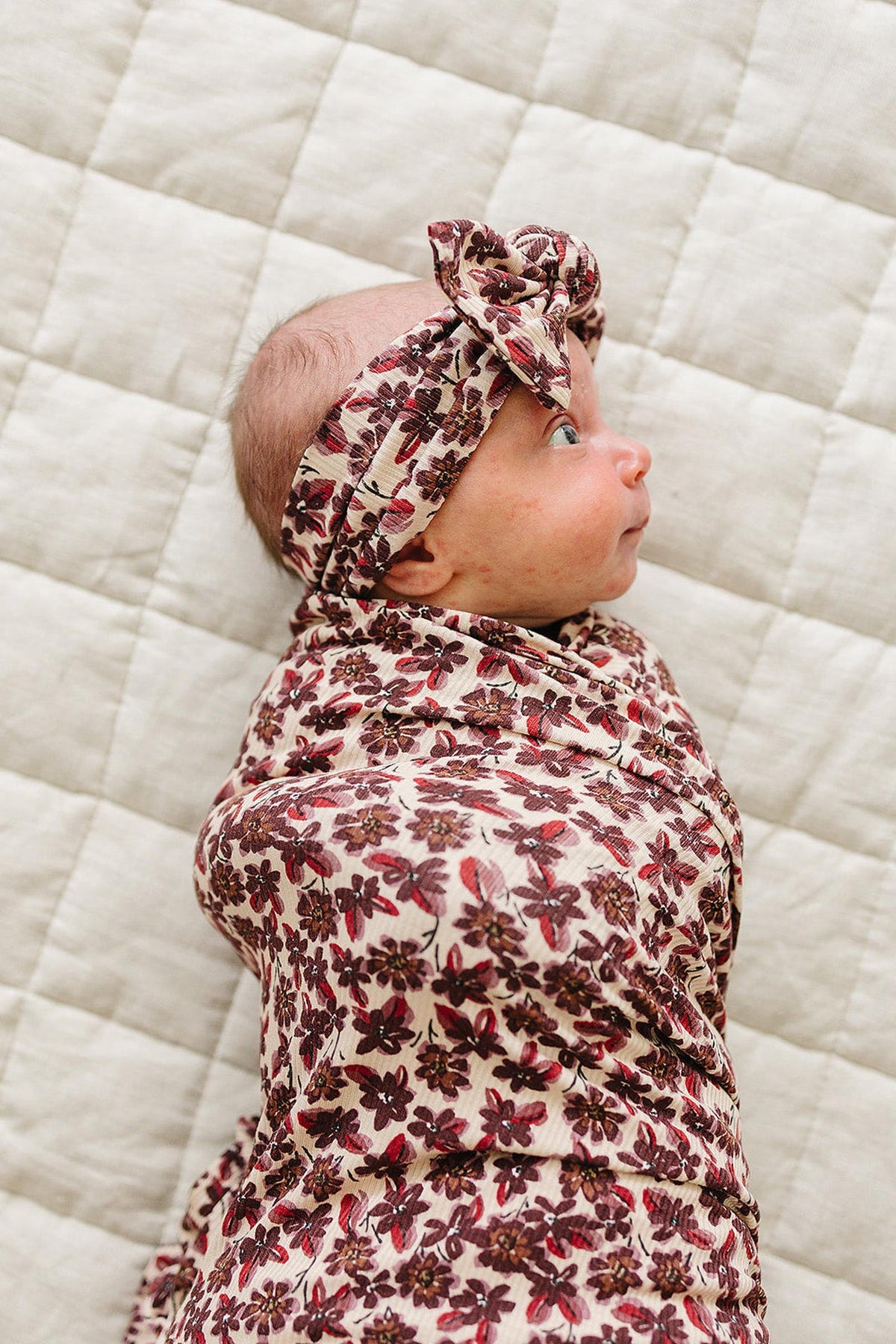 A baby swaddled in an antique floral ribbed bamboo blanket, wearing a matching headband. The soft, breathable fabric features a delicate floral pattern, resting on a textured, light-colored quilt.