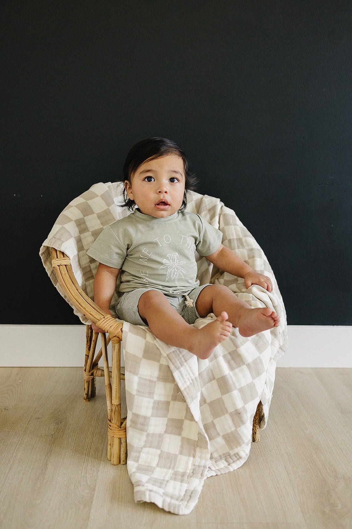 A toddler wearing a green Take Me To The Beach t-shirt sits in a wicker chair with a plaid blanket.