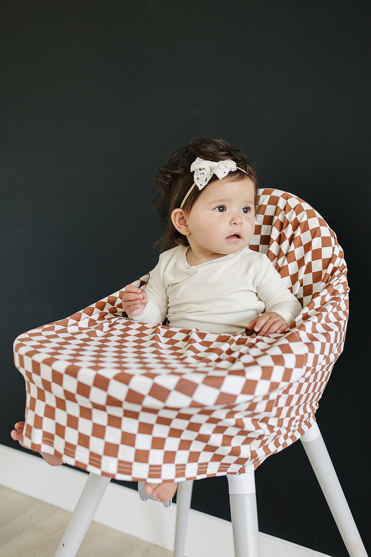 A baby sits in a high chair covered with a rust checkered bamboo fabric, wearing a white outfit.