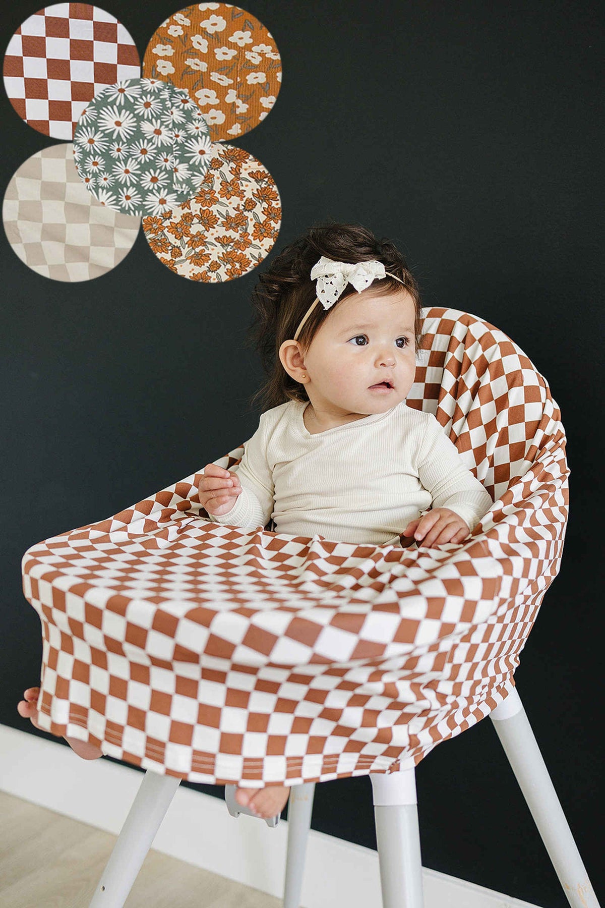 A baby sits in a high chair covered with a brown and white checkered fabric, wearing a cream outfit.