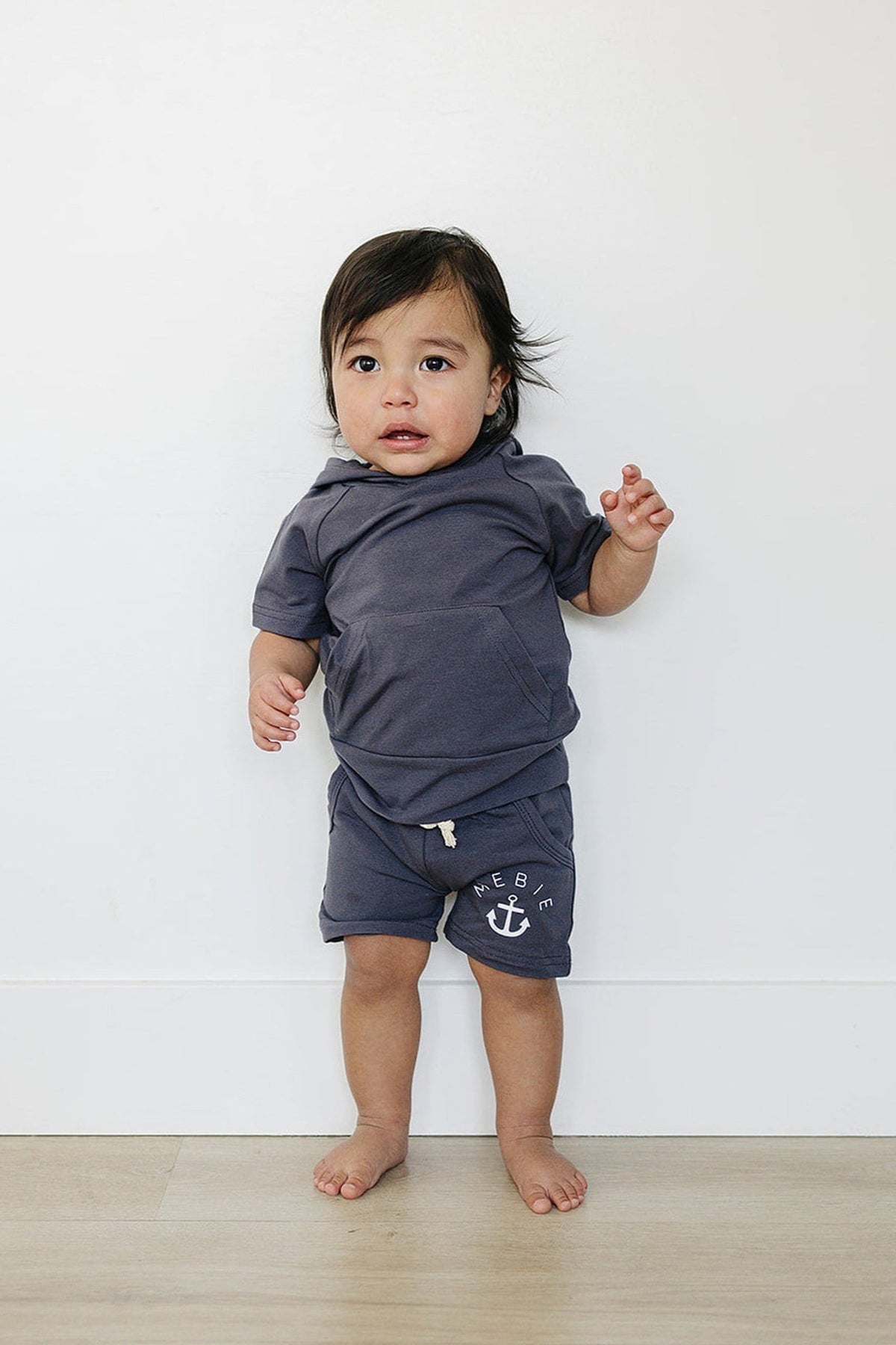 A toddler in a slate hooded T-shirt and shorts stands against a white wall on a wooden floor.