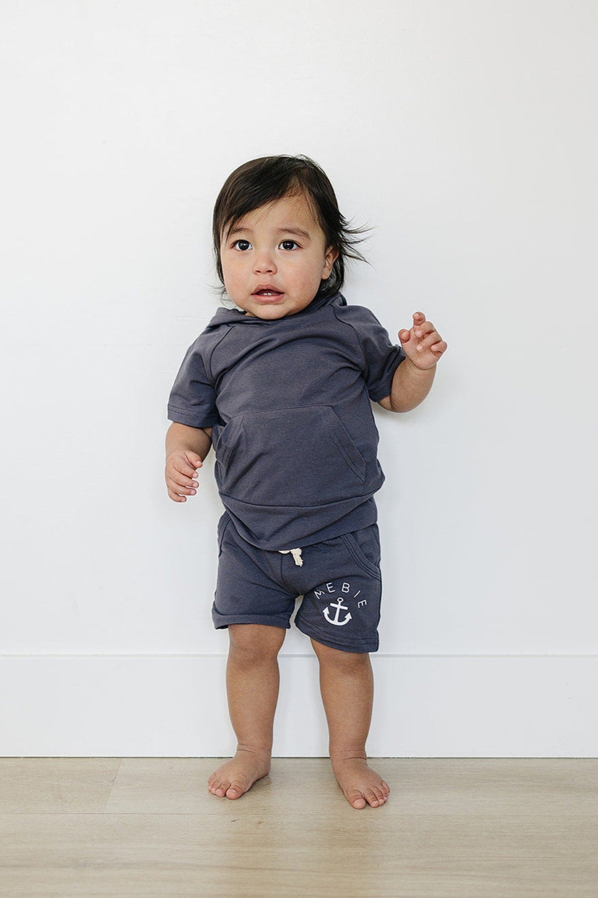 A toddler wearing a slate-colored cotton outfit with Mebie branding and anchor detail stands against a white background.