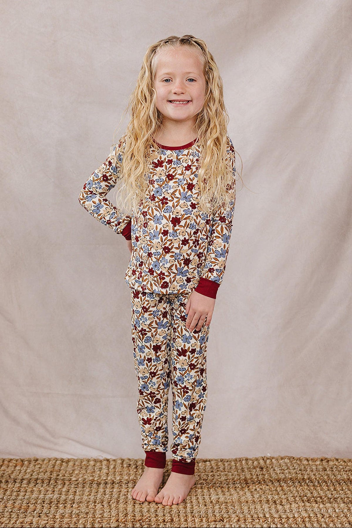 A smiling child wearing a floral-patterned, ribbed bamboo cozy set with maroon cuffs stands on a woven rug.