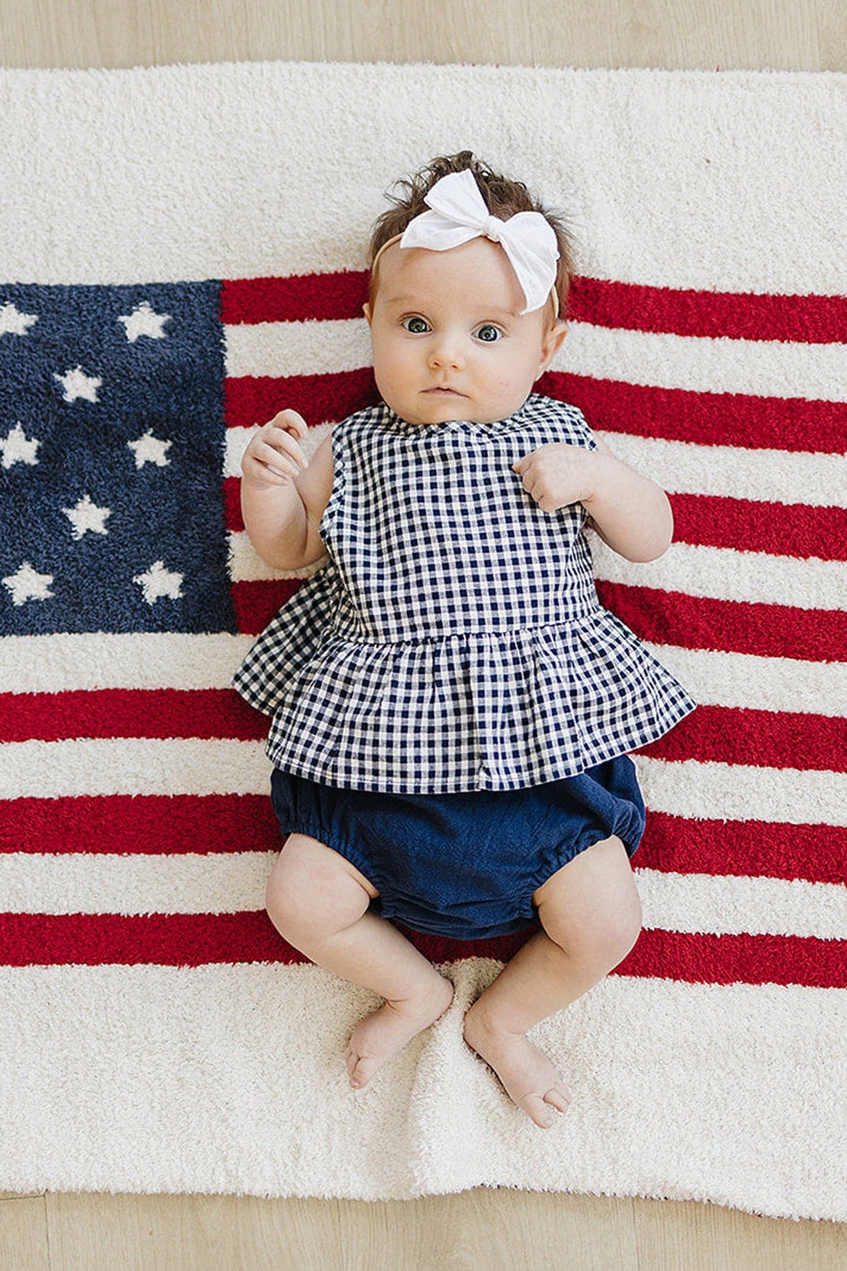 A baby wearing a navy gingham peplum set with bloomers lies on an American flag-themed rug.