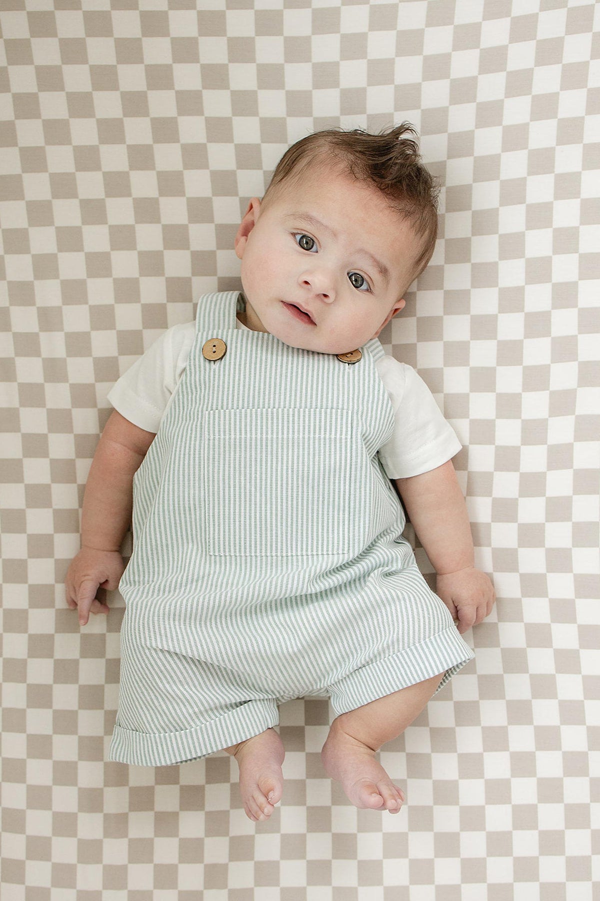A baby in striped overalls lies on a taupe checkered crib sheet, looking upwards.