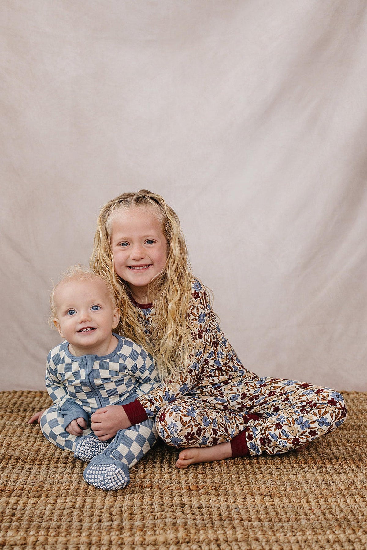 Two children sit on a woven rug, wearing checkered and floral patterned pajamas, smiling against a neutral backdrop.