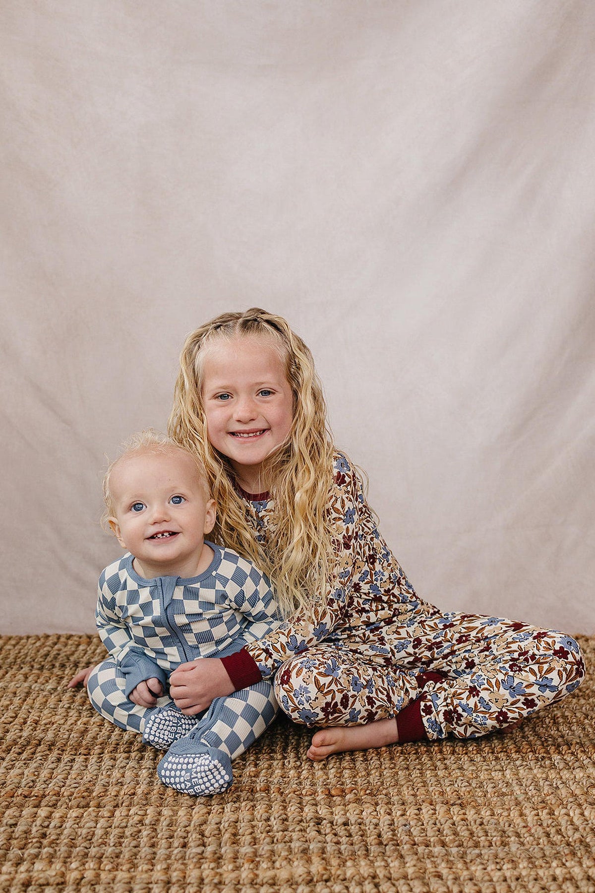 Two smiling children sit on a woven rug, wearing patterned pajamas; one in blue checkered, the other in floral.