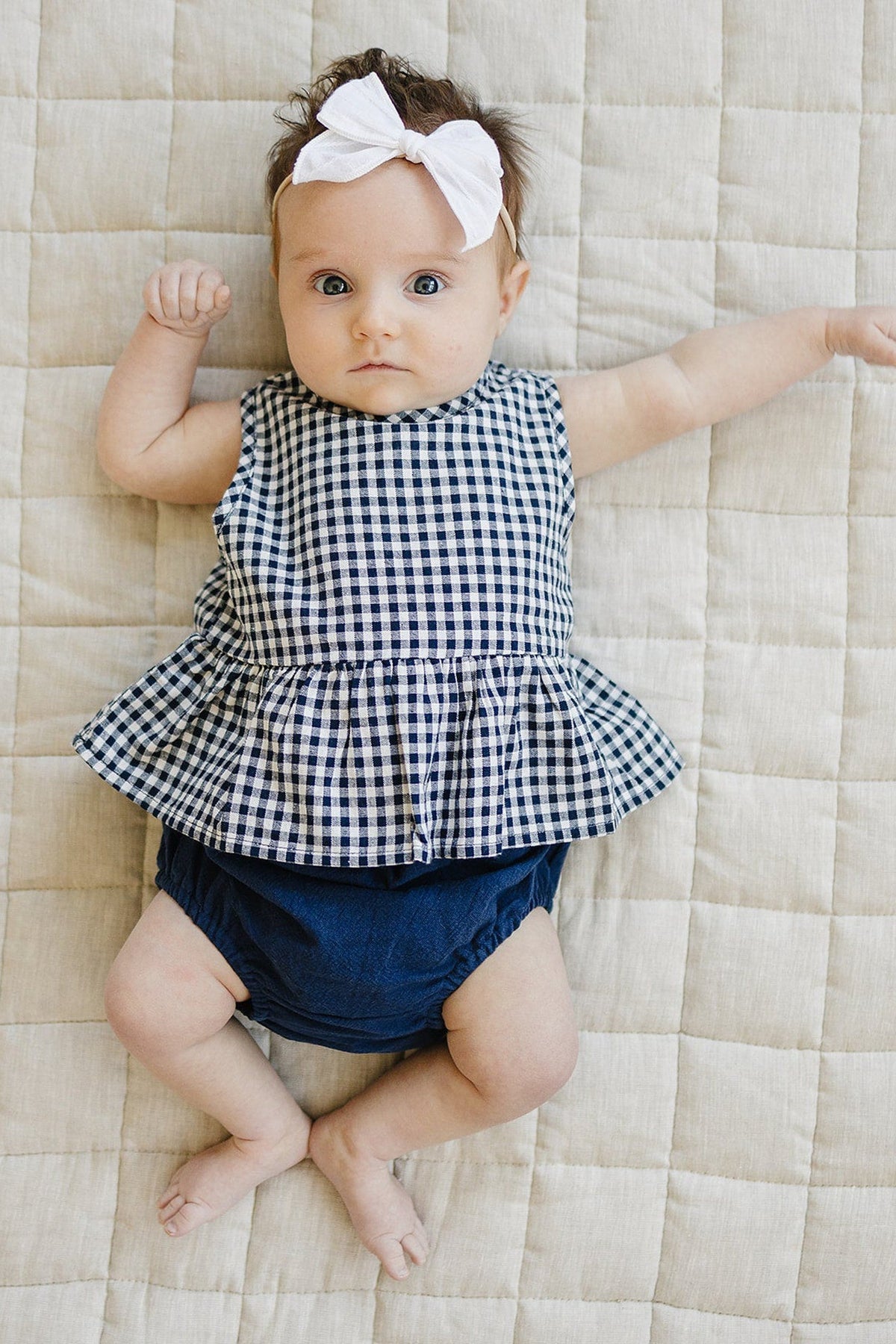 Baby wearing a navy gingham peplum top and bloomers, lying on a quilted beige blanket, with a white bow.