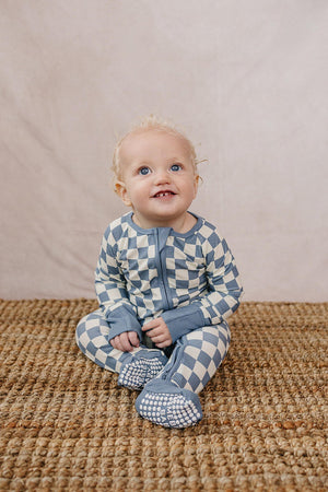 A baby wearing blue checkered pajamas sits on a woven mat, smiling against a neutral background.
