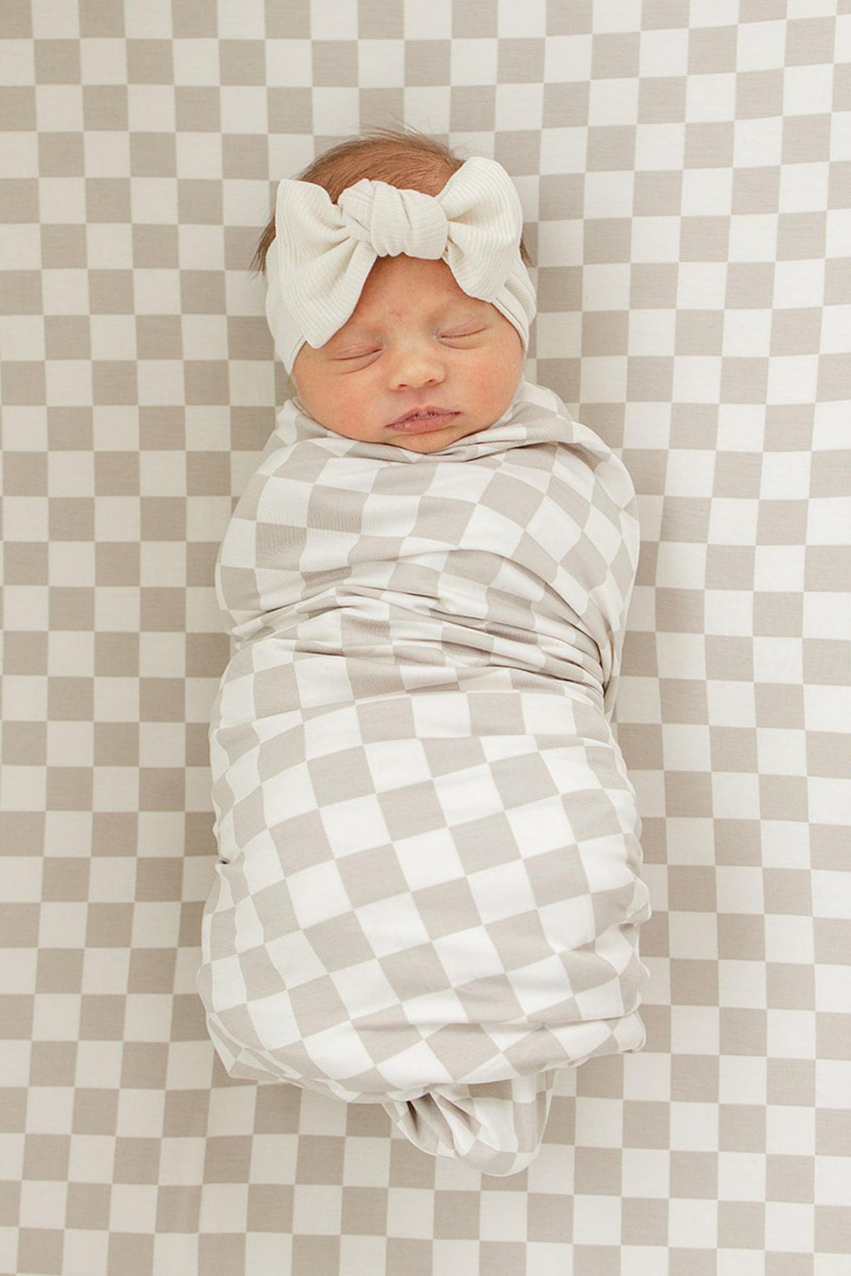 A baby wrapped in a taupe checkered swaddle with a matching headband, lying on a checkered crib sheet.