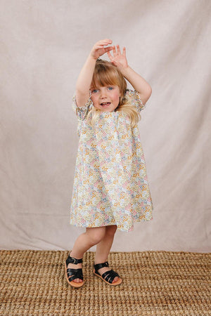 A young girl in a cheerful floral dress with pastel colors poses playfully against a neutral backdrop. The dress features short sleeves and a relaxed fit, perfect for summer adventures.
