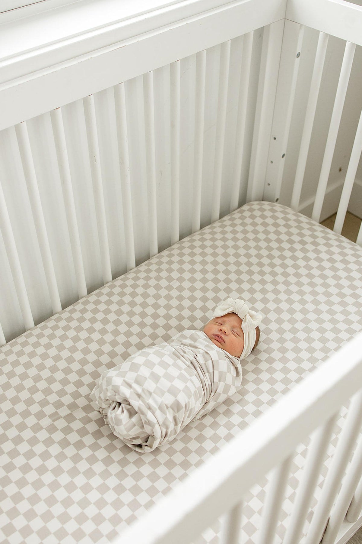 A baby is swaddled in a checkered blanket on a taupe checkered crib sheet in a white crib.
