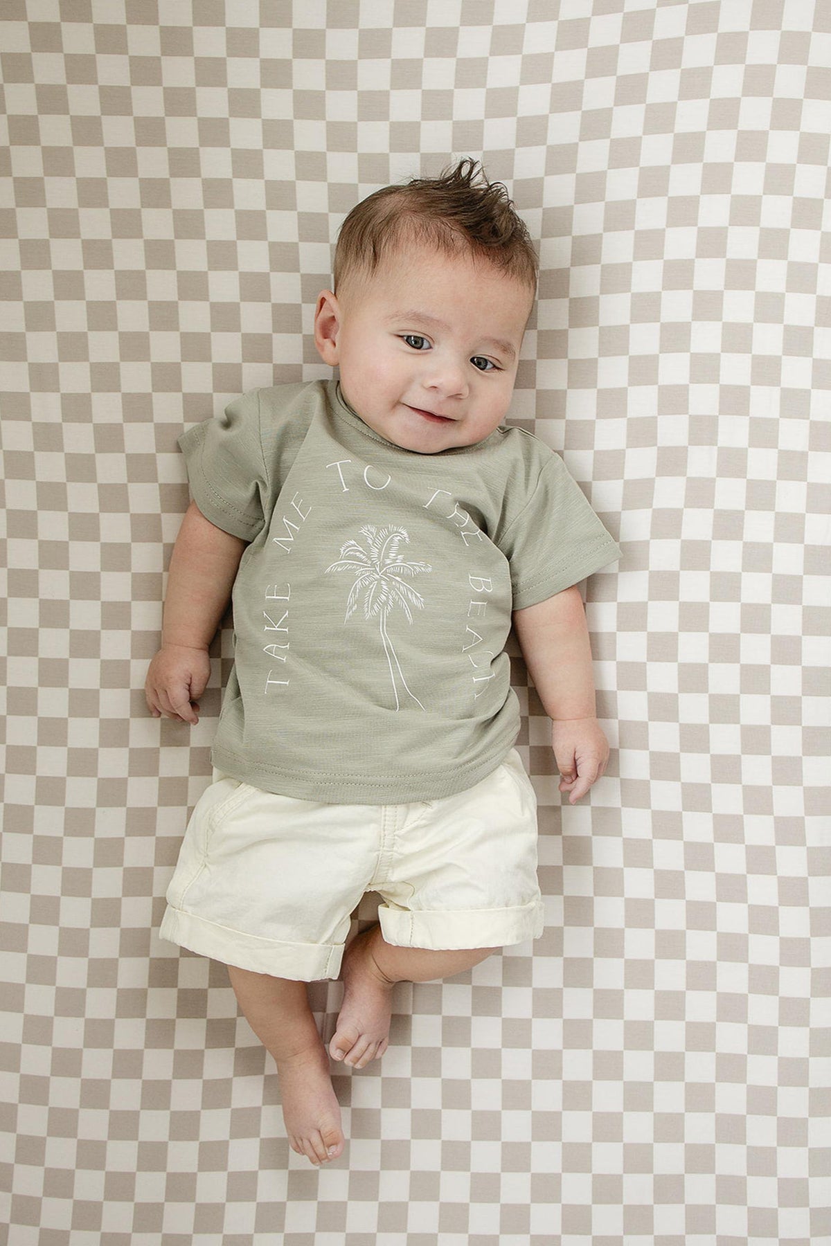 A baby lies on a taupe checkered crib sheet, wearing a green shirt and cream shorts.