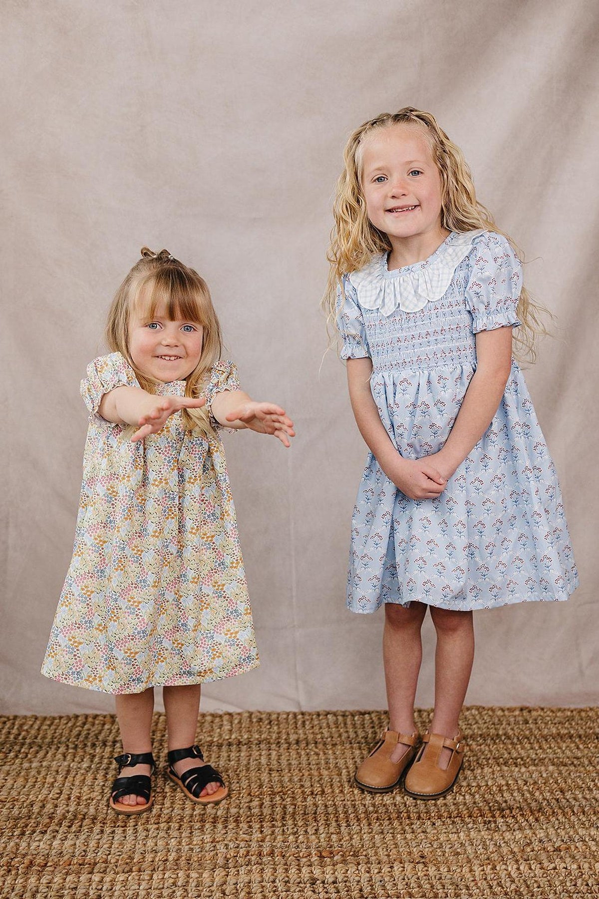 Two young girls stand on a textured rug, one wearing a floral cotton dress and the other in a light blue dress with a white collar. Both display cheerful smiles.