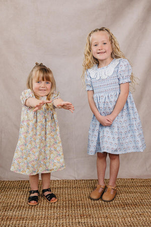 Two young girls stand on a textured rug, one wearing a floral cotton dress and the other in a light blue dress with a white collar. Both display cheerful smiles.