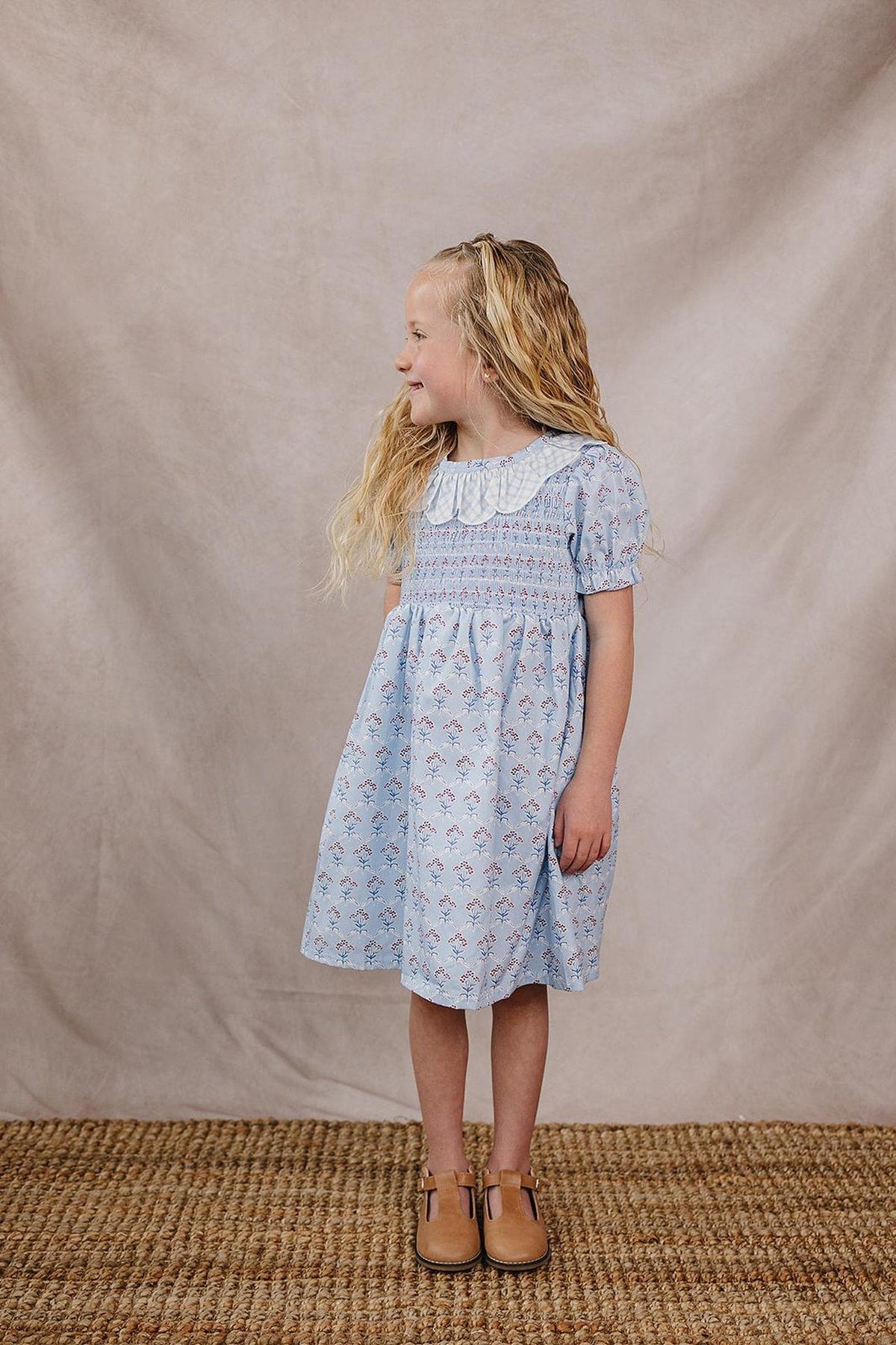A young girl in a light blue dress with a gingham collar and floral accents stands on a natural woven rug against a soft beige backdrop, smiling and ready for.
