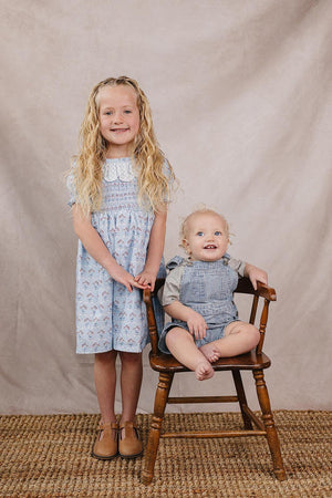 A smiling girl in a light blue floral dress stands beside a toddler in a gray and white checkered romper, both seated on a wooden chair against a neutral backdrop.