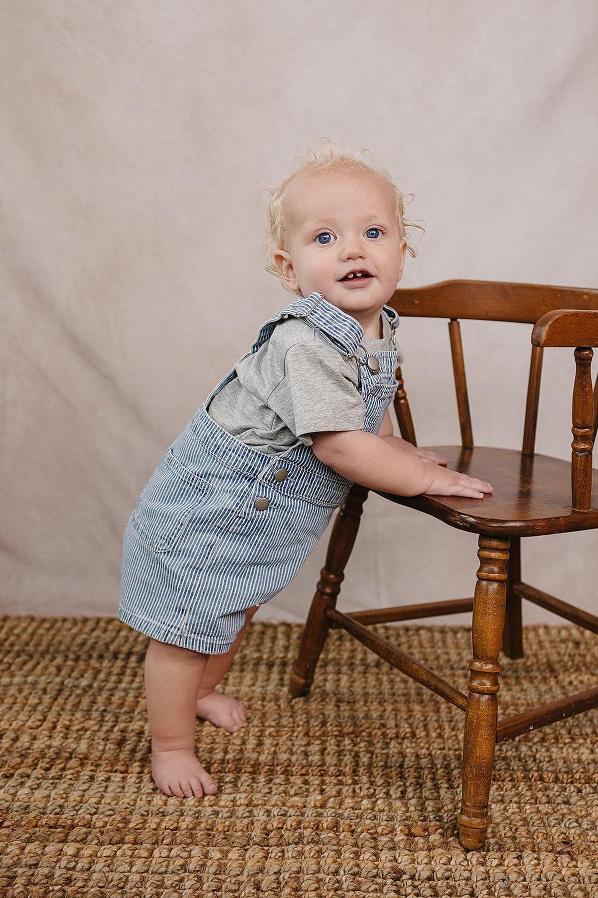 A toddler with curly hair wears a grey t-shirt and blue-striped overalls, leaning on a wooden chair against a neutral.