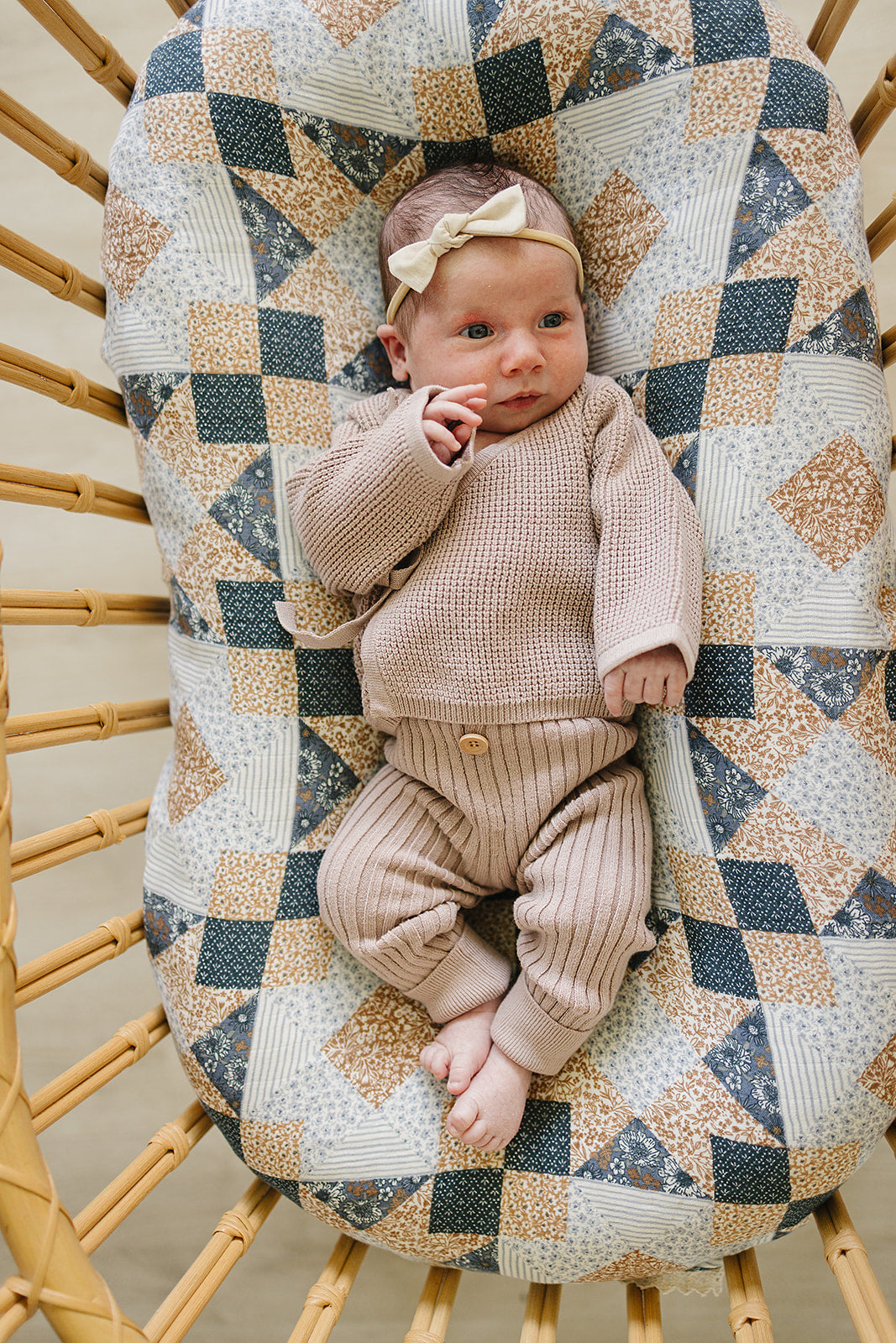 A baby comfortably rests on a patterned fabric with a diamond design, wearing a soft mauve knit cardigan and matching ribbed leggings, complemented by a cute bow headband.
