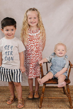 Three children are posed together, wearing a Sea Air Sun cotton t-shirt, a patterned dress, and striped overalls.