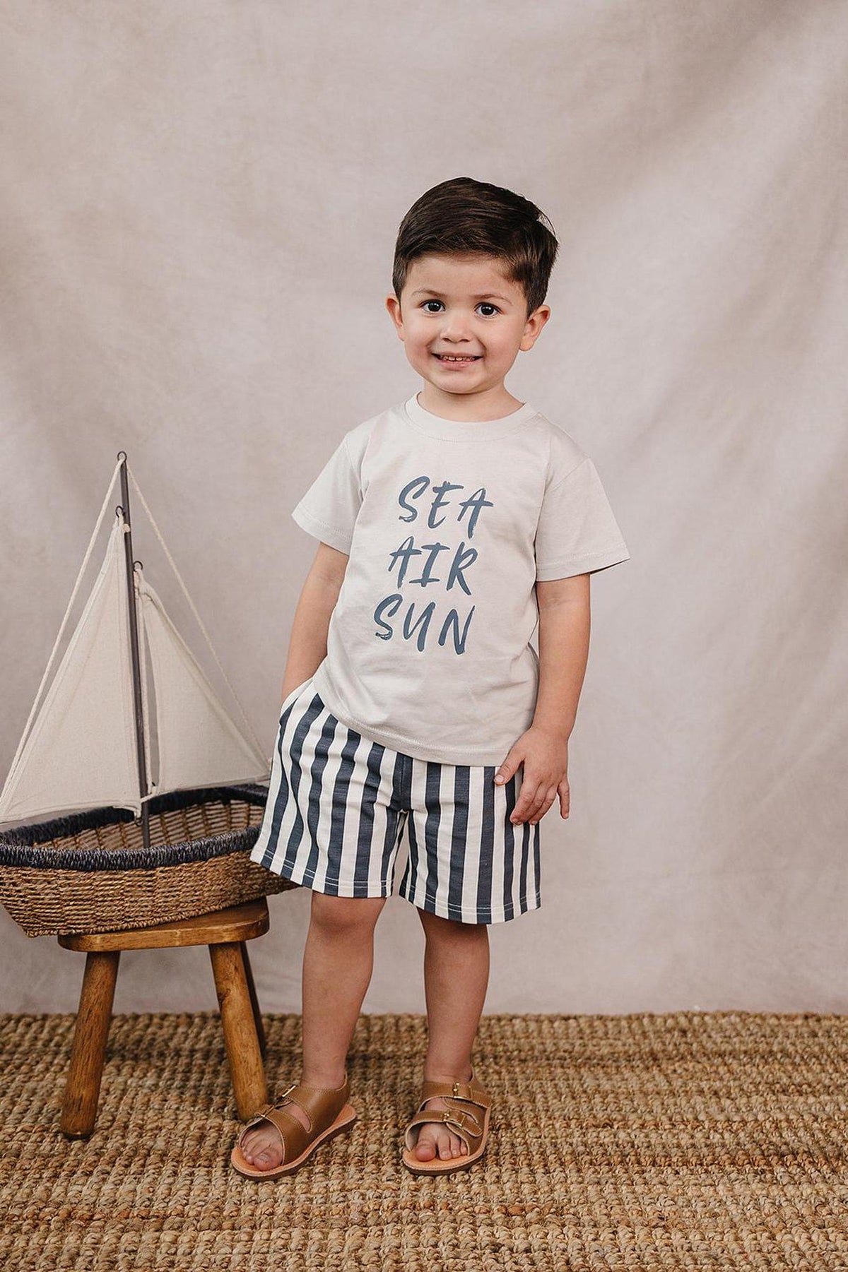 A smiling child wears a beige SEA AIR SUN cotton t-shirt with navy striped shorts, standing next to a decorative.