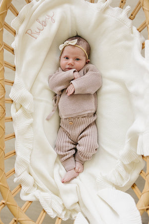 A baby lies peacefully in a woven bassinet, wrapped in a soft, cream-colored blanket, wearing a mauve knit outfit with ribbed leggings and a matching bow headband.