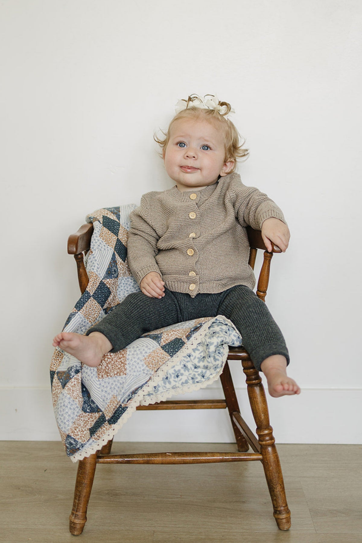 A toddler sits on a wooden chair, wearing a cozy brown knit cardigan with buttons and grey leggings. A patterned quilt drapes over the chair, enhancing the warm, inviting scene.