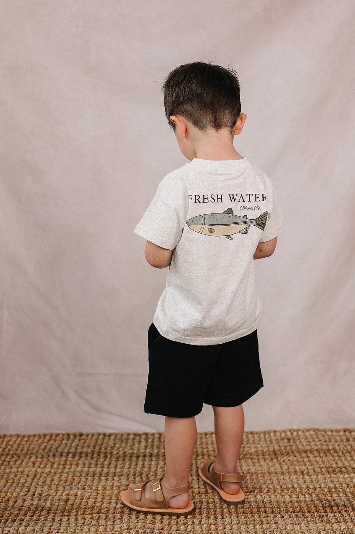 A child wearing a white T-shirt with a fish graphic and the words Fresh Water, standing on a woven rug.