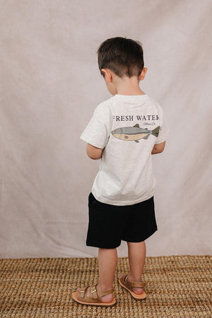 A child wearing a white T-shirt with a fish graphic and the words Fresh Water, standing on a woven rug.