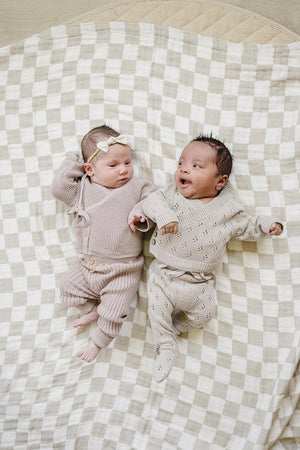 Two infants lie on a soft, checkered blanket, dressed in coordinated pointelle knit outfits in soft taupe and beige, showcasing delicate details and cozy comfort.