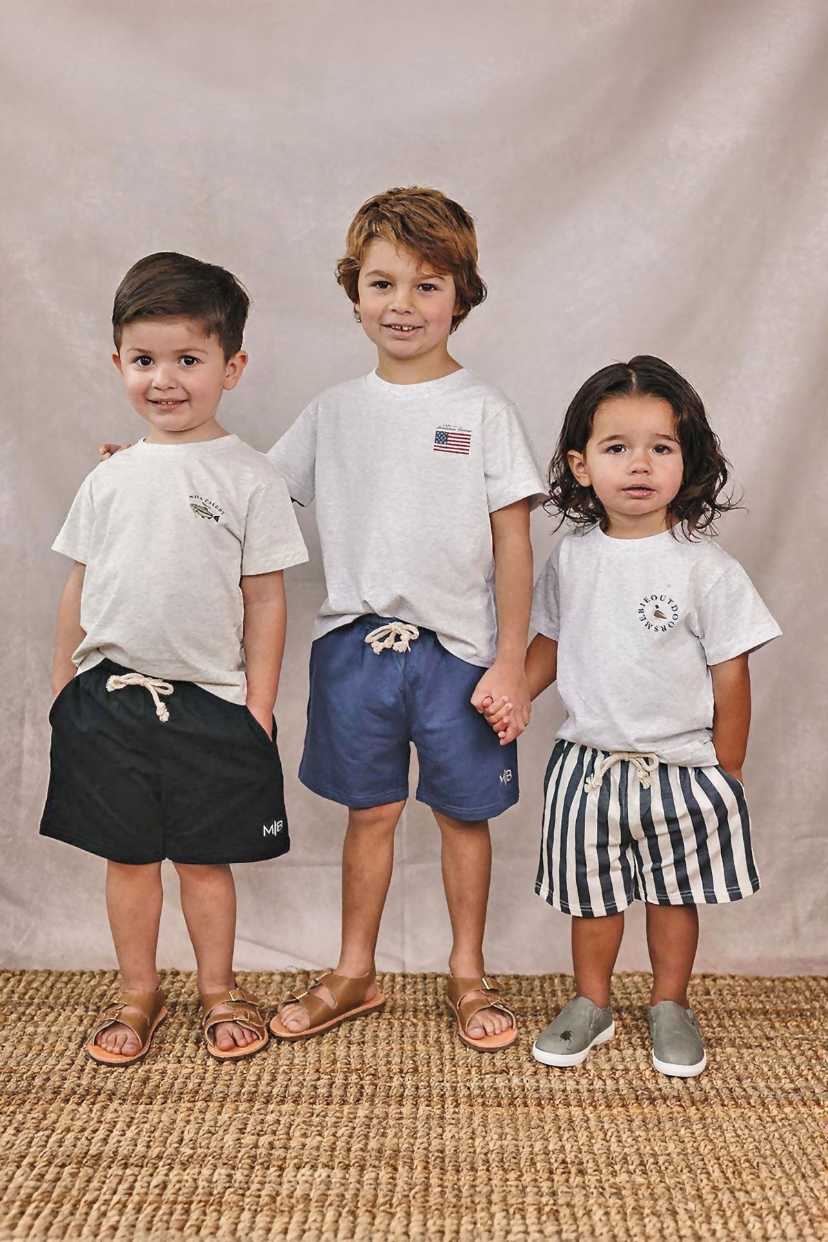 Three children wearing white t-shirts and shorts stand on a woven rug, showcasing casual, comfortable summer outfits.