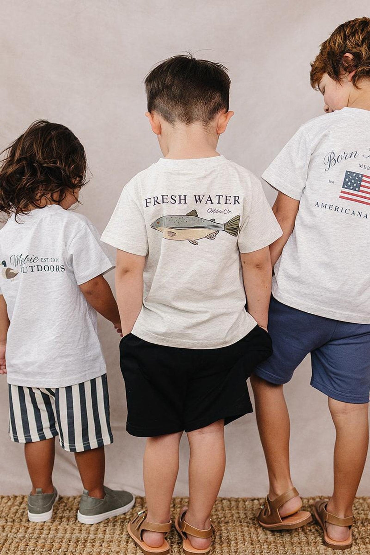 Three children wearing Mebie Baby cotton T-shirts with outdoor-themed graphics stand together on a woven rug.