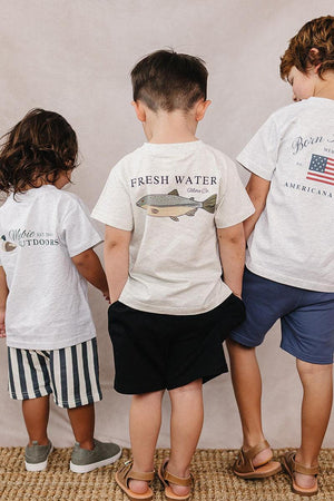 Three children wearing Mebie Baby cotton T-shirts with outdoor-themed graphics stand together on a woven rug.
