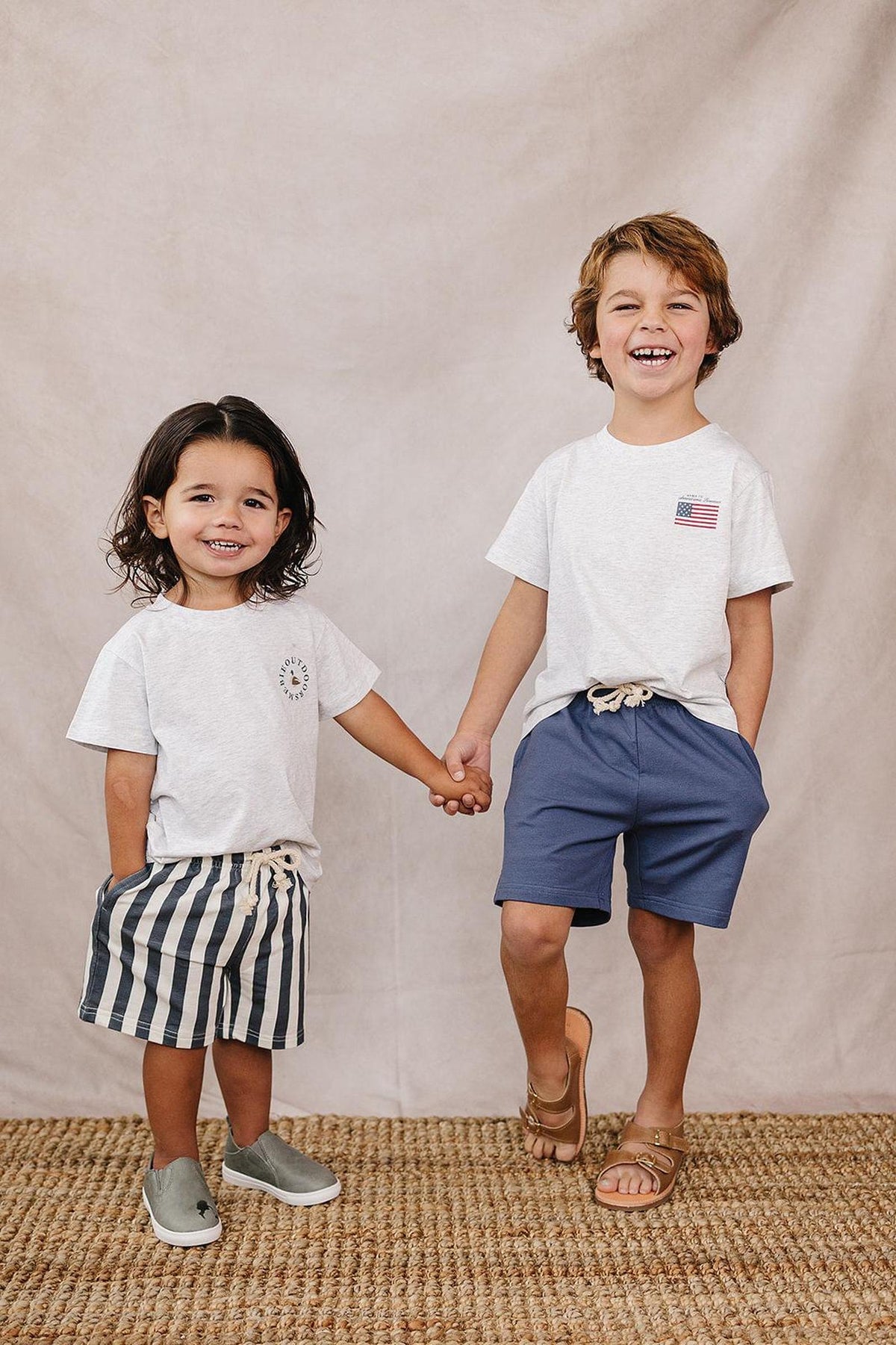 Two smiling children holding hands, wearing light gray t-shirts; one with striped shorts, the other with blue shorts.