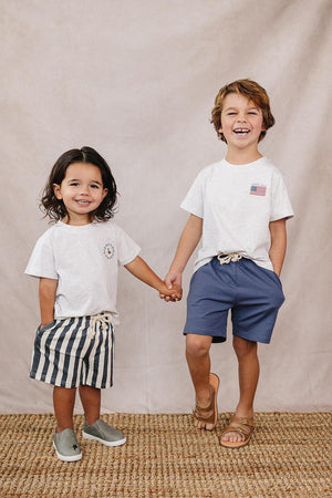 Two smiling children holding hands, wearing light gray t-shirts; one with striped shorts, the other with blue shorts.