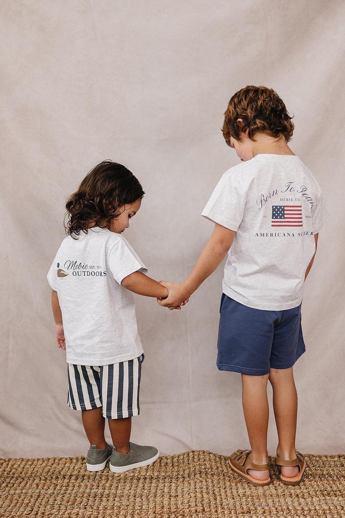 Two children holding hands, wearing Mebie Baby Americana T-shirts, one with striped shorts, standing on a woven rug.