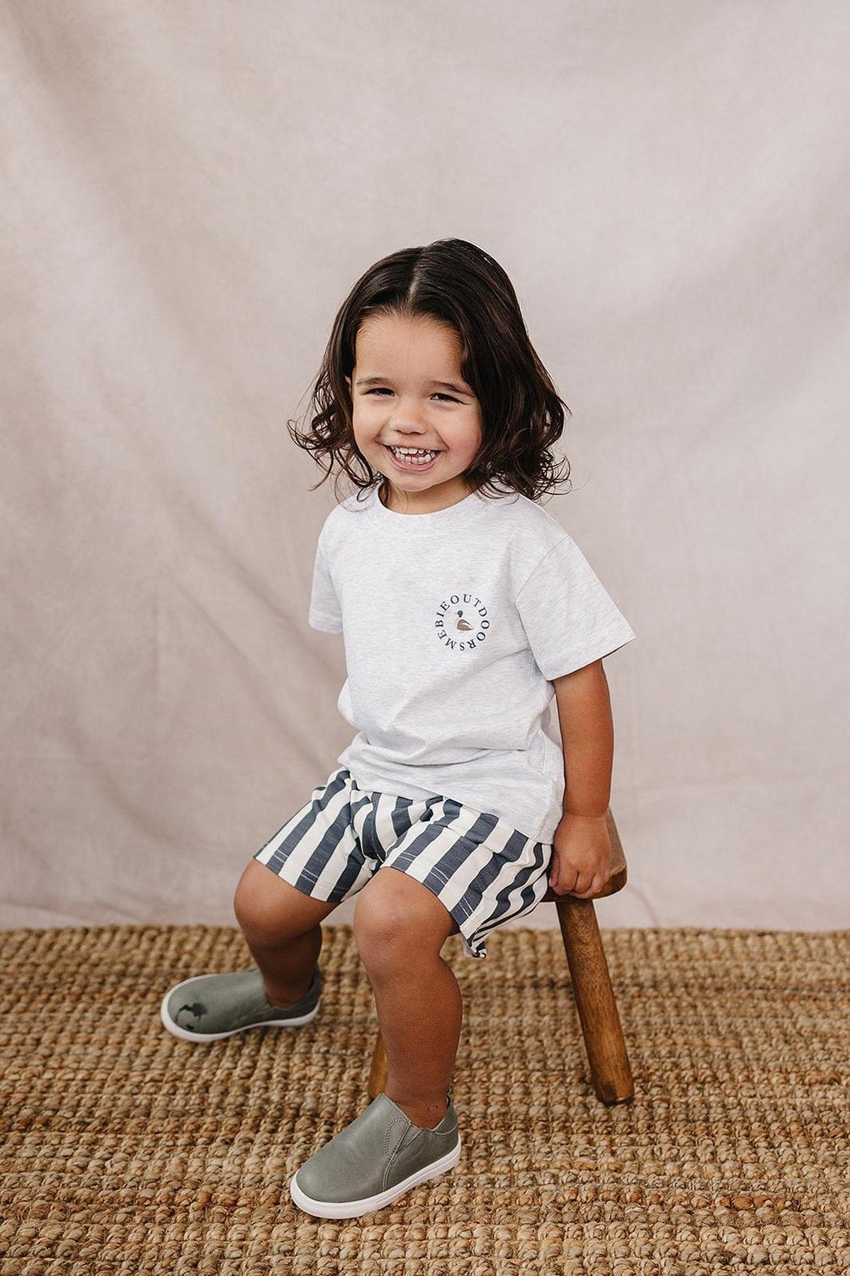 A smiling child wearing a light gray Mebie Outdoors T-shirt, striped shorts, and gray slip-on shoes sits on a stool.