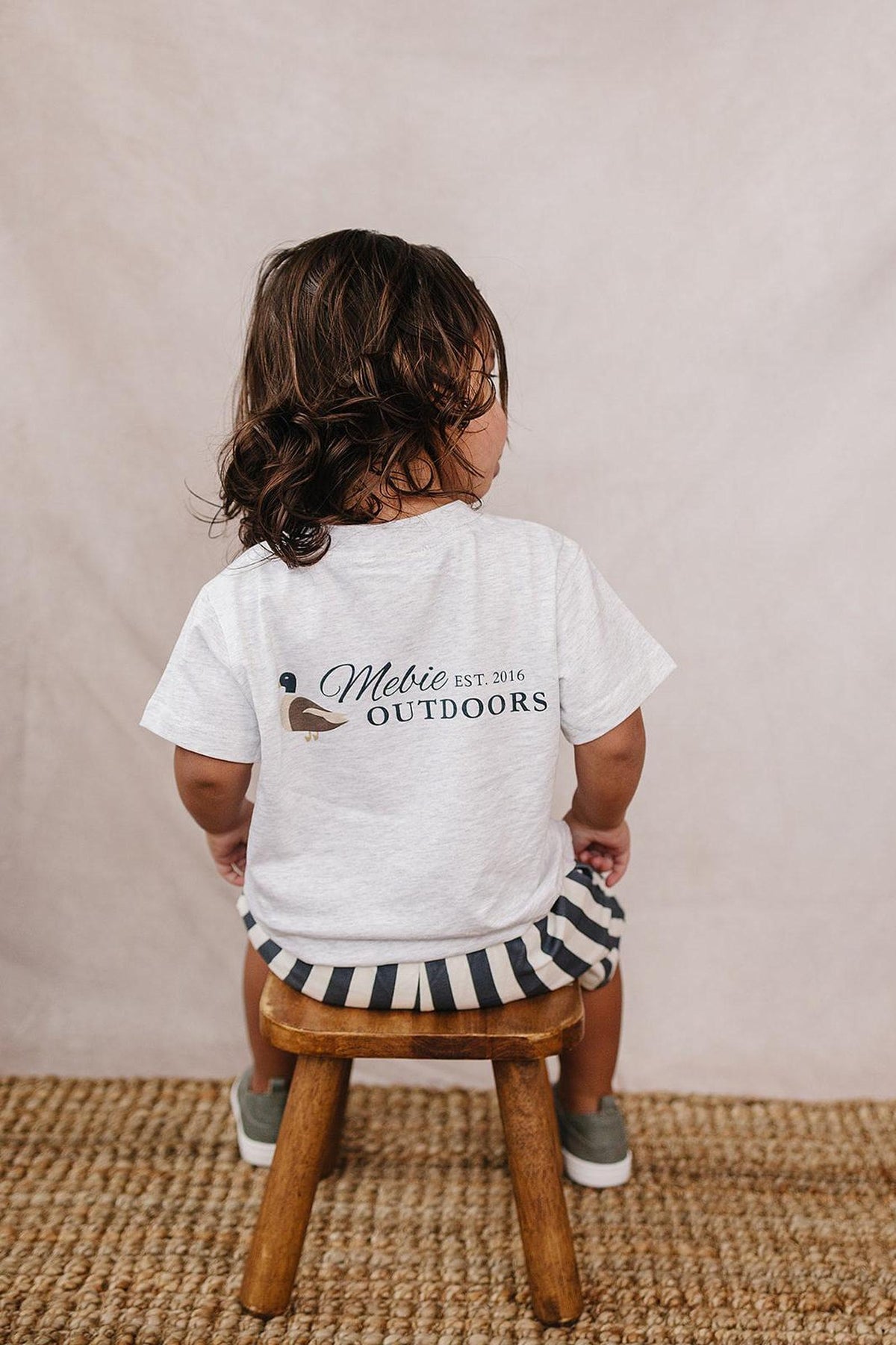 A child wearing a white Mebie Outdoors T-shirt with a duck graphic sits on a wooden stool.